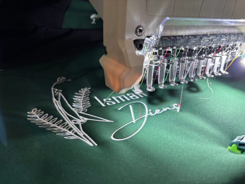 Stickmaschine stickt einen Schriftzug mit Ornament auf grünen Stoff
